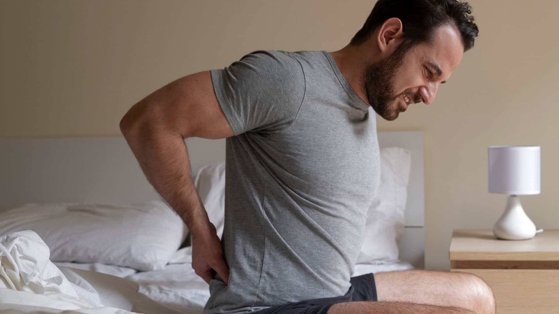 A man wincing and holding his lower back as he sits on the edge of his bed.