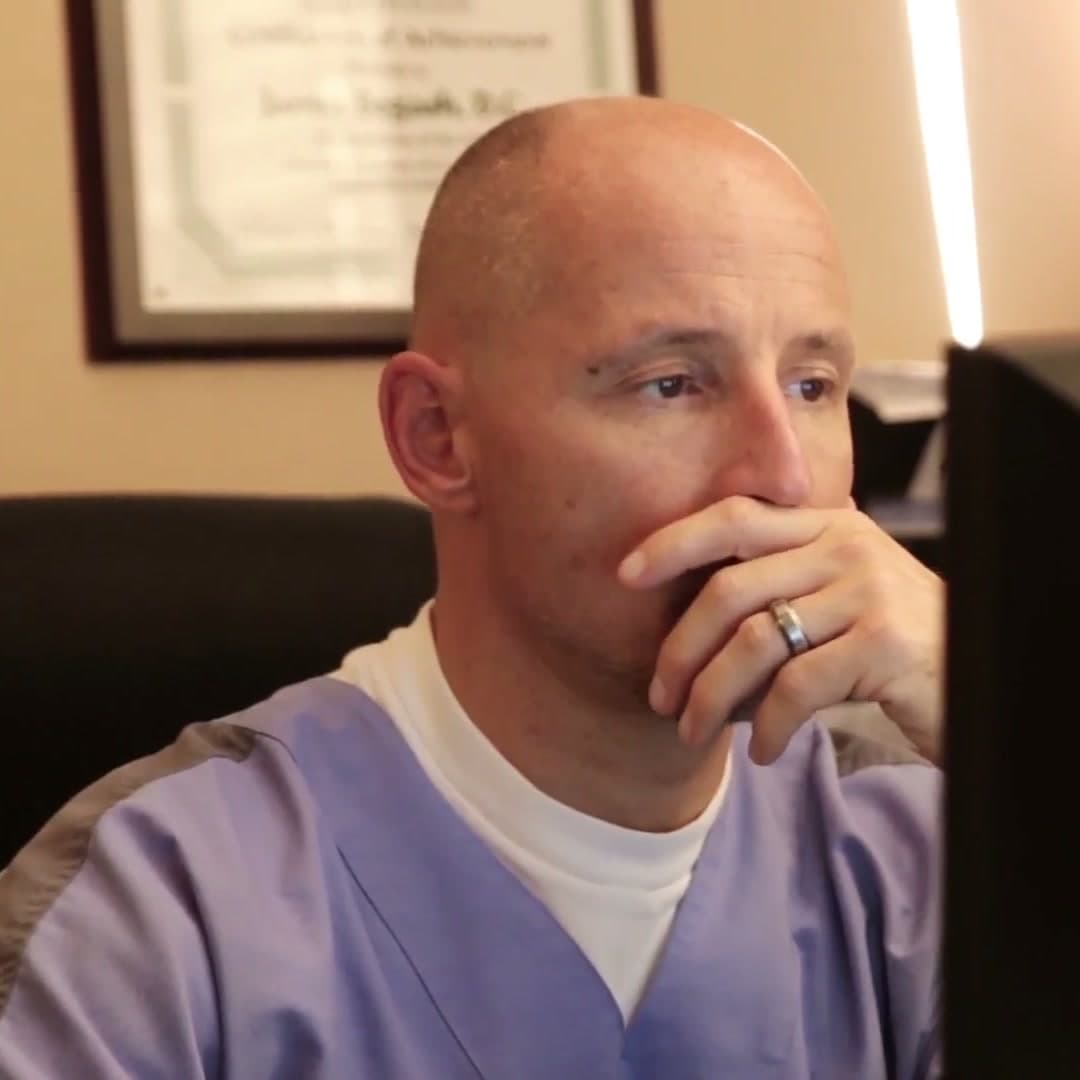 Dr. Bogash at his desk reading on screen, his framed certificate on the wall behind him.